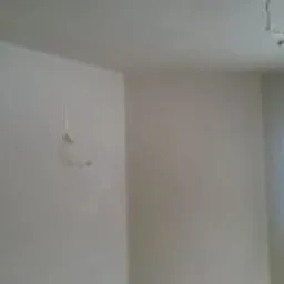 Wnętrze pomieszczenia po nałożeniu świeżego, jasnego tynku. Widoczne gładkie ściany i sufit, zarys okna w tle, oraz drobne niedoskonałości w strukturze tynku na ścianie po lewej stronie.